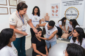 Alunas em treinamento prático de doulagem no curso Doula Brasil, com banner ao fundo mencionando a regulamentação da lei 3.946/2021 e carga horária de 120 horas.