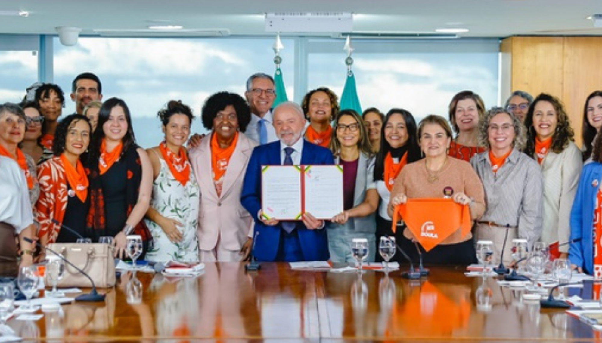 Presidente Lula sorrindo ao lado de doulas e ministros, segurando o documento sancionado da Lei das Doulas no Palácio do Planalto.