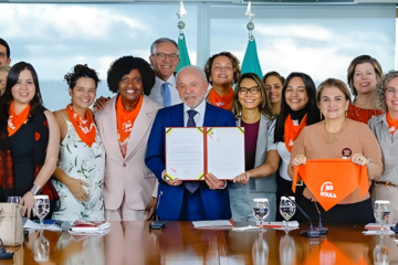 Presidente Lula sorrindo ao lado de doulas e ministros, segurando o documento sancionado da Lei das Doulas.